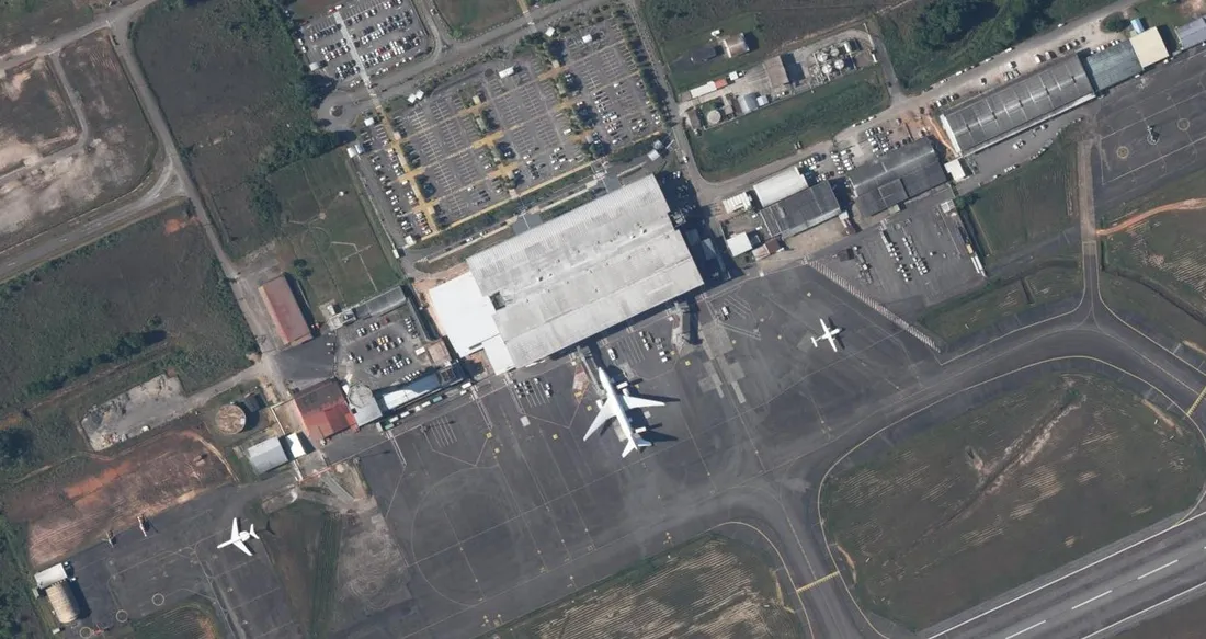 Vue aérienne verticale de l'aérogare de Cayenne – Félix-Éboué à Matoury : le long bâtiment à toiture métallique, un gros-porteur au poste de stationnement devant la salle d'embarquement, un bimoteur régional sur l'aire voisine et les parkings de l'aérogare en haut à gauche