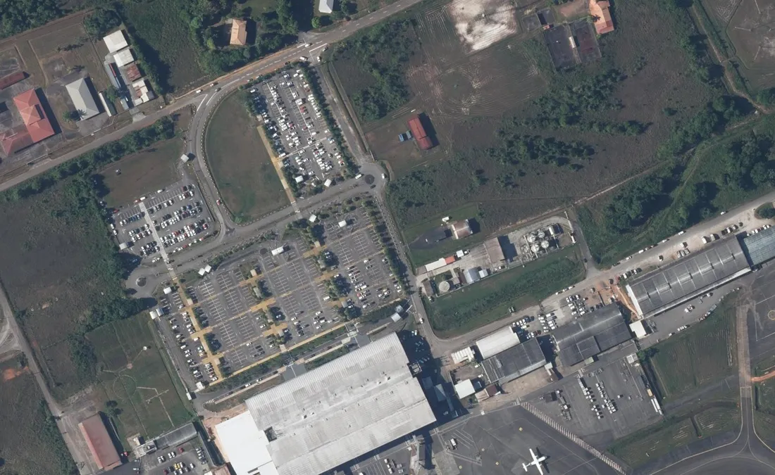 Vue aérienne des parkings de l'aéroport Félix-Éboué à Matoury : trois nappes de stationnement séparées par des rangées d'arbres, la voie de desserte en boucle qui vient de la route de Rochambeau, et la toiture de l'aérogare dans l'angle inférieur