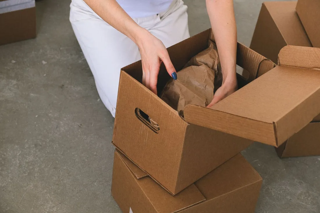 Femme agenouillée emballant des affaires enveloppées dans un carton de déménagement, entourée d'autres cartons prêts à partir