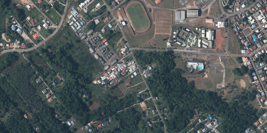 Orthophotographie aérienne verticale du bourg de Matoury, en Guyane : maisons individuelles et petits collectifs de part et d'autre de la route, stade, piscine municipale et groupe scolaire, au contact du massif boisé du mont Grand Matoury