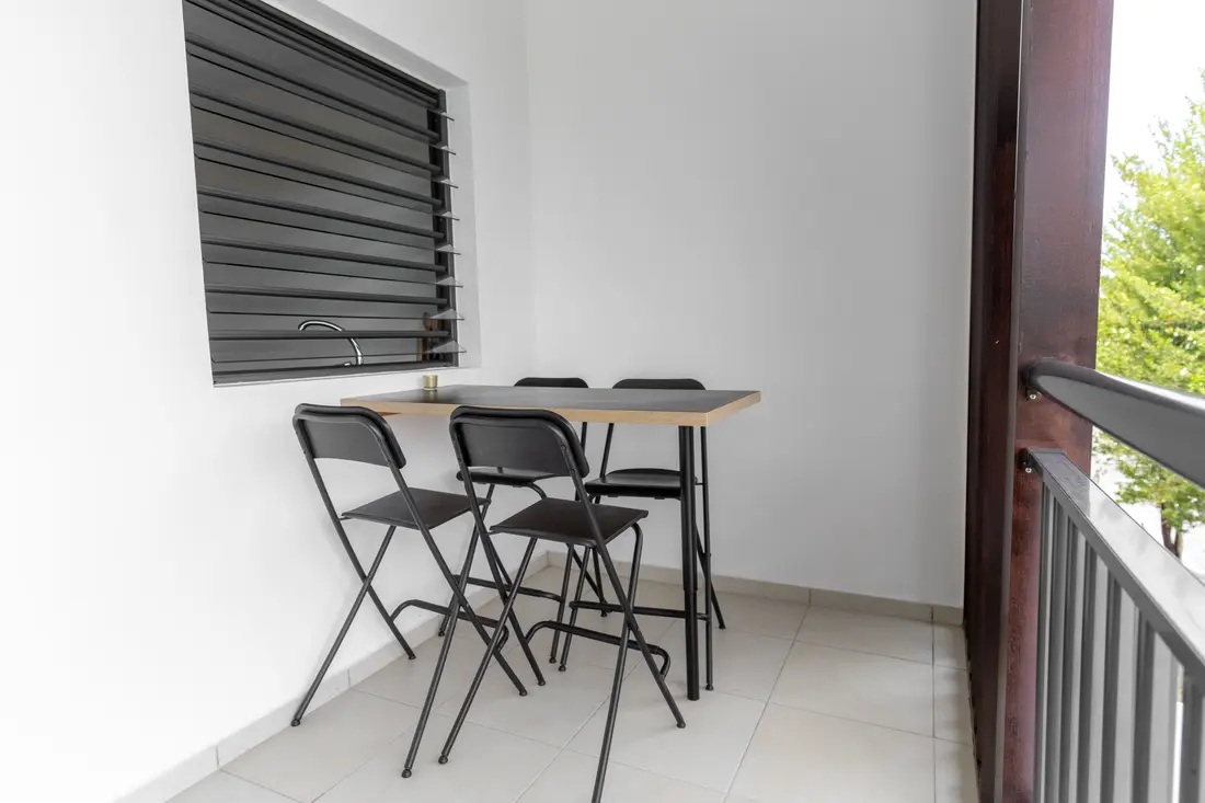 Balcon du Nid de Thalys : table haute noire et quatre chaises pliantes devant une jalousie, arbre en contrebas