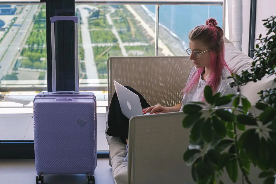Femme en déplacement professionnel travaillant sur son ordinateur portable dans un logement lumineux, valise cabine posée devant la baie vitrée