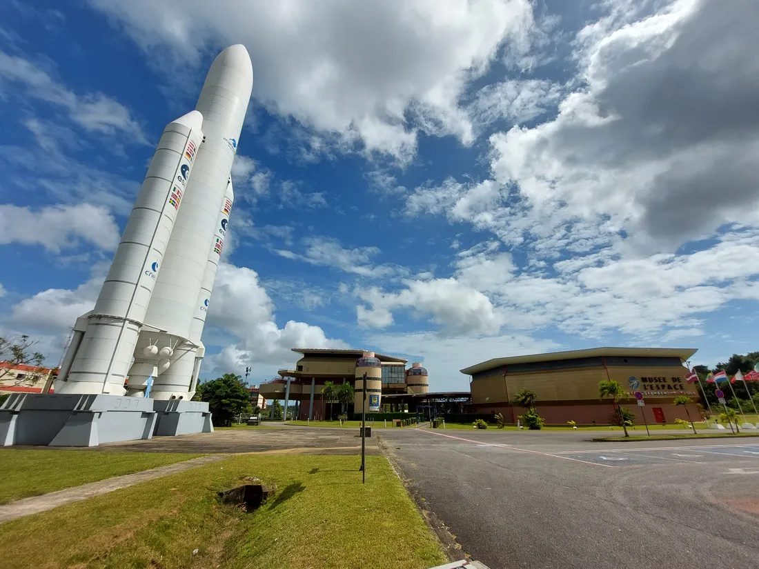 L'accueil du Centre spatial guyanais à Kourou : à gauche la maquette grandeur nature d'Ariane 5 dressée sur son socle, au centre le bâtiment d'accueil du CSG, à droite le hall à toiture ocre du musée de l'Espace, devant un parking et une pelouse déserts