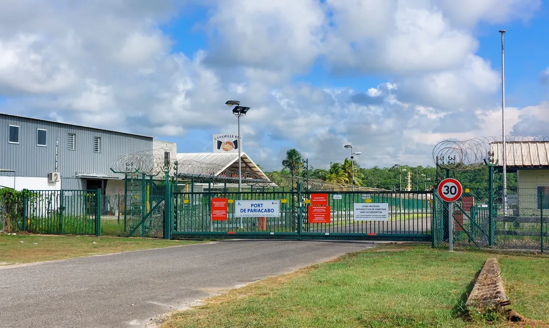 Le portail fermé du port de Pariacabo à Kourou : grille métallique verte surmontée de barbelés, panneau bleu « Port de Pariacabo », panneau de limitation à 30 km/h, hangar à bardage gris à gauche et halle couverte au fond