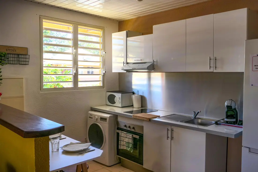Cuisine d’un appartement Cayenne Chic : plaques, four, lave-linge, micro-ondes et réfrigérateur sous les placards blancs