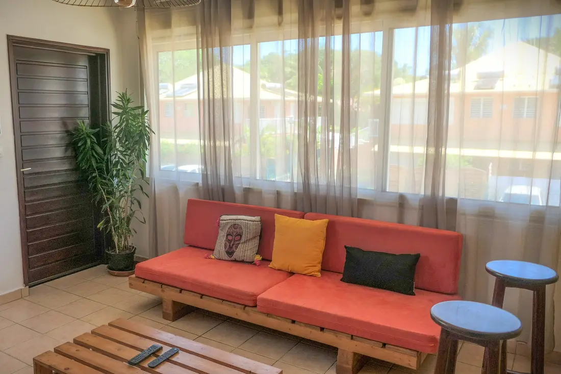 Séjour d’un appartement Cayenne Chic : canapé corail sur base en bois de palette, grandes baies voilées donnant sur la résidence