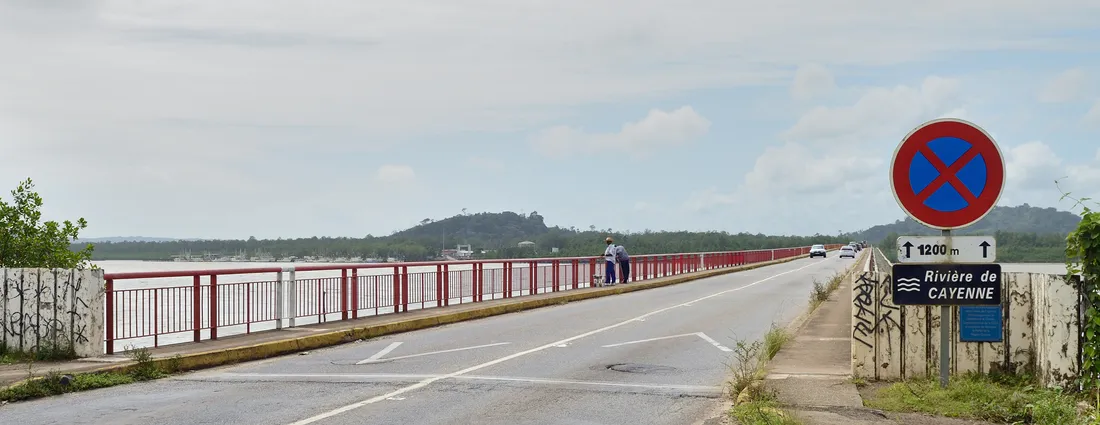Le pont du Larivot sur la RN1 en Guyane, vu depuis la chaussée : une voie par sens, garde-corps rouge, panneau « Rivière de Cayenne 1200 m » à droite, mâts de voiliers au fond à gauche