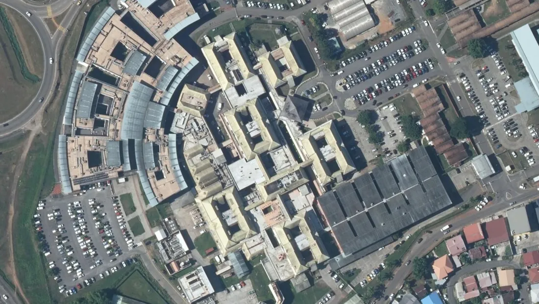 Vue aérienne verticale du site hospitalier Andrée-Rosemon à Cayenne : corps de bâtiments en peigne à toitures claires au centre, long bâtiment en arc de cercle couvert d'une galerie vitrée à l'ouest, vastes parkings remplis de voitures, voie rapide et bretelle d'accès en bordure gauche