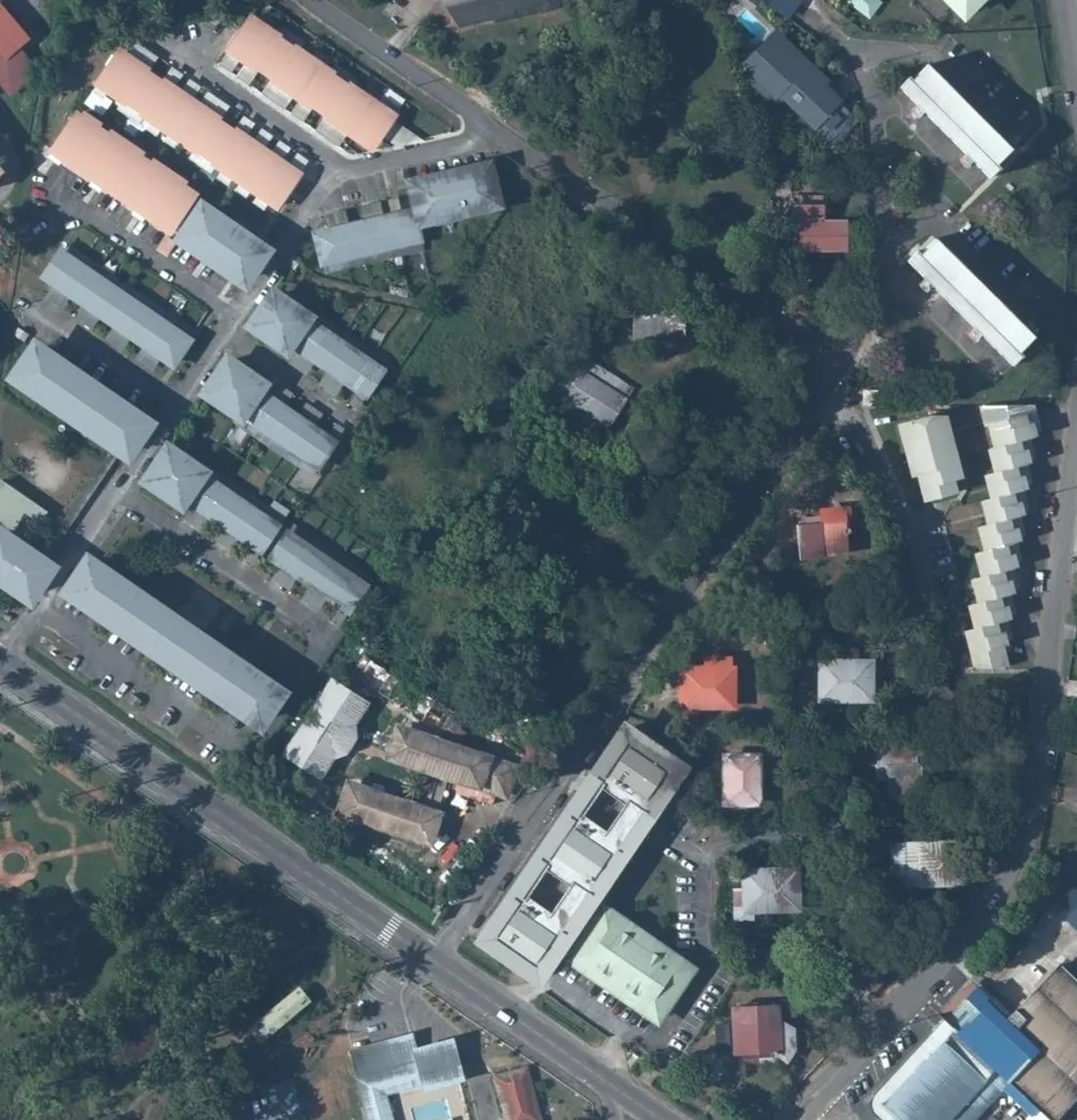 Vue aérienne verticale du terrain d'assiette de la future cité judiciaire de Cayenne, quartier Rebard : une grande parcelle boisée et non bâtie occupe le centre de l'image, bordée à gauche par les barres de logements collectifs et leurs parkings, en bas par l'avenue du Général Virgile et un bâtiment administratif allongé, à droite par les maisons individuelles de la Cité Rebard