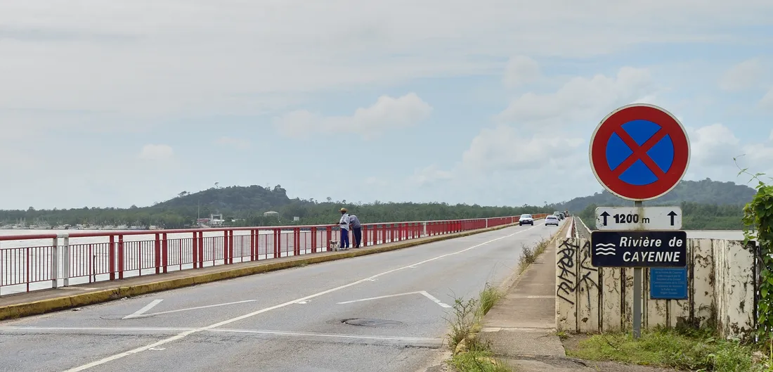 Le pont du Larivot sur la RN1 en Guyane, vu depuis le tablier : une chaussée à une voie par sens bordée à gauche d'un garde-corps métallique rouge et blanc, deux piétons accoudés à la rambarde, quelques voitures au loin, l'estuaire de la rivière de Cayenne et un ponton de pirogues à gauche, la colline boisée du Larivot au fond, un panneau bleu « Rivière de Cayenne » et un panneau d'interdiction de stationner à droite