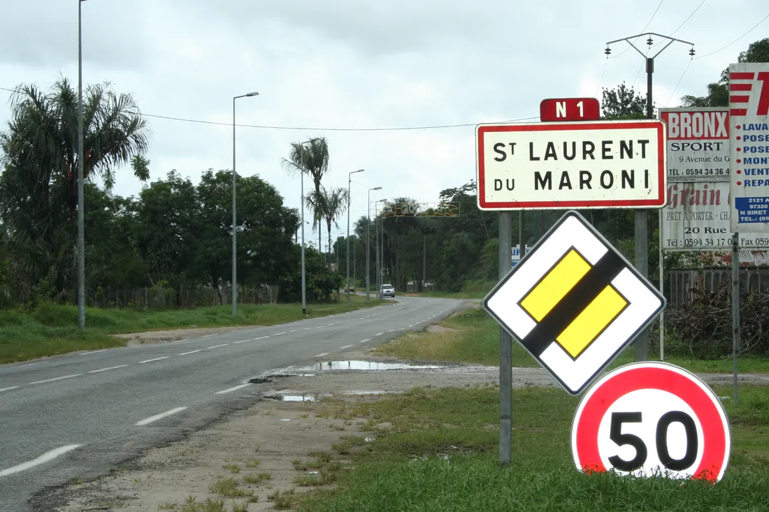 Panneau d'entrée d'agglomération « St Laurent du Maroni » surmonté de la cocarde rouge « N 1 », planté au bord de la RN1 en Guyane : une chaussée à deux voies bordée d'éclairage public et de palmiers, un panneau de route prioritaire et une limitation à 50 km/h au premier plan, quelques enseignes commerciales et un pylône électrique sur la droite