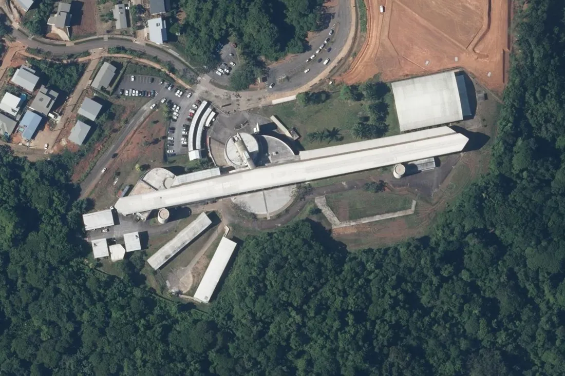 Vue aérienne verticale du lycée Léon Gontran Damas à Rémire-Montjoly : long bâtiment linéaire blanc adossé au relief boisé, gymnase à toiture sombre et parking de cars scolaires