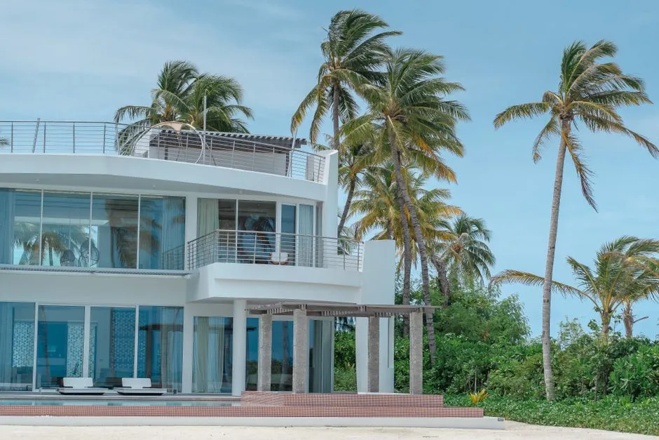 Villa contemporaine blanche en bord de plage entouree de cocotiers sous un ciel bleu, location de vacances face a la mer