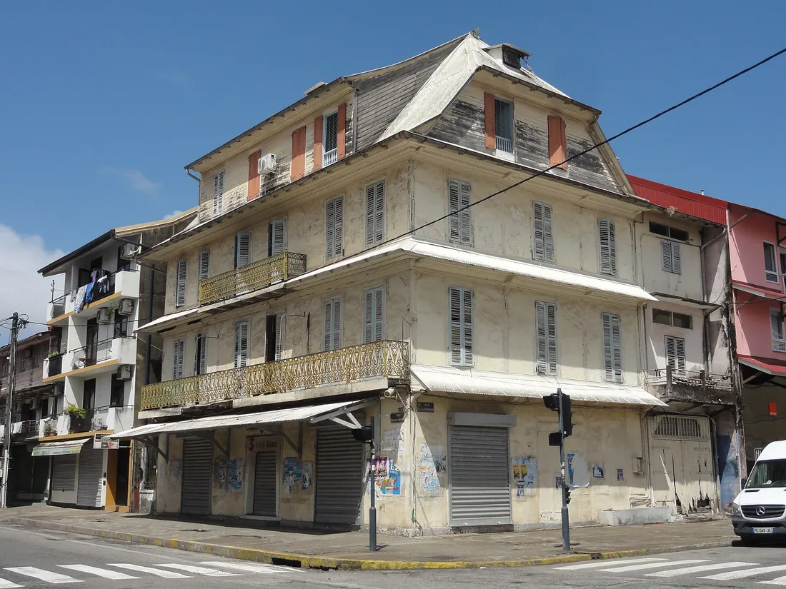 L'immeuble Vitalo à Cayenne, à l'angle des rues François-Arago et Camille-Bernard : façade décrépie, persiennes closes à tous les étages et rideaux métalliques baissés au rez-de-chaussée.