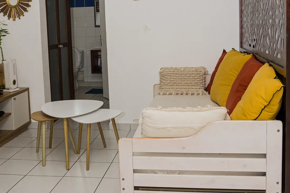 Séjour du Cocon Cayennais : banquette en bois clair à coussins jaunes et terracotta, tables gigognes blanches, salle d’eau en arrière-plan