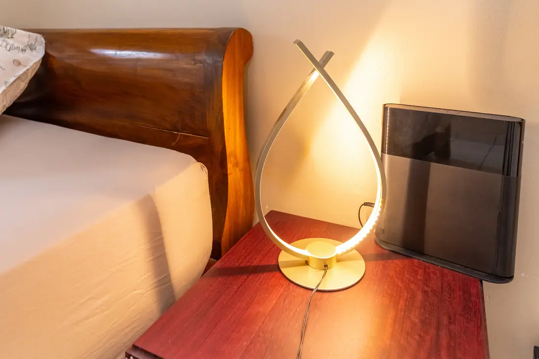 Chevet en bois rouge du T2 Mahury : lampe dorée en goutte allumée près de la tête de lit en bois massif