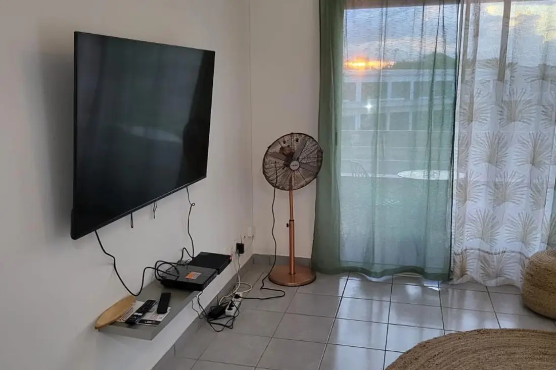 Séjour de Yana Chic au coucher du soleil : grande télévision murale, ventilateur sur pied cuivré et baie vitrée donnant sur la terrasse