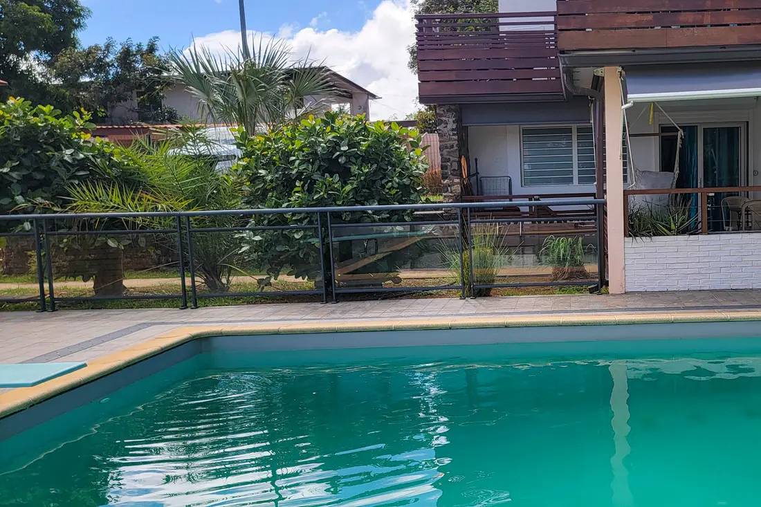 Piscine de la résidence Sweet Dreams à Cayenne : bassin au premier plan, terrasse couverte et façade en bois derrière la clôture vitrée