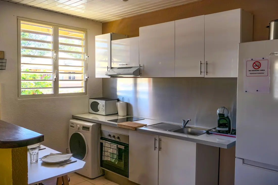 Cuisine équipée d’un appartement Cayenne Chic : meubles blancs, crédence inox, plaques, four, lave-linge et réfrigérateur