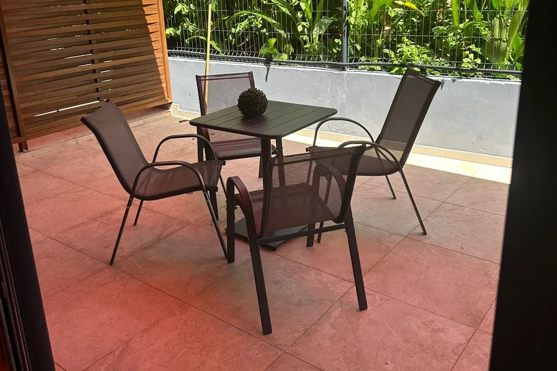 Terrasse d’un logement de la Résidence Francis : table carrée et quatre chaises sous un auvent rouge, verdure dense derrière la clôture