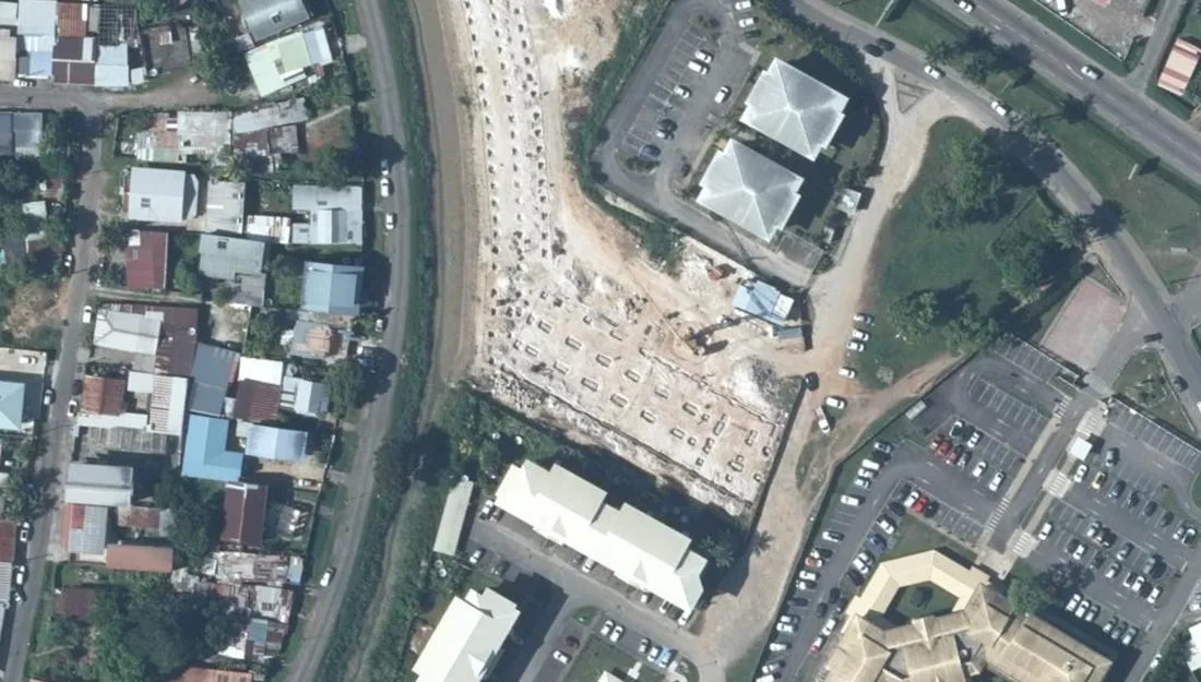 Vue aérienne verticale de la parcelle du 250 route de Baduel à Cayenne : terrain en chantier avec plots de fondation, parkings et bâtiments à toitures pyramidales, bordé à l'ouest par le quartier pavillonnaire de Baduel