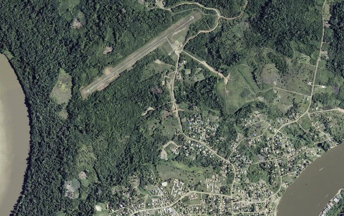 Vue aérienne verticale de Maripasoula en Guyane : piste d'atterrissage en latérite au milieu de la forêt, bourg dispersé le long de pistes de terre, et fleuve Maroni bordant l'image à gauche et à droite