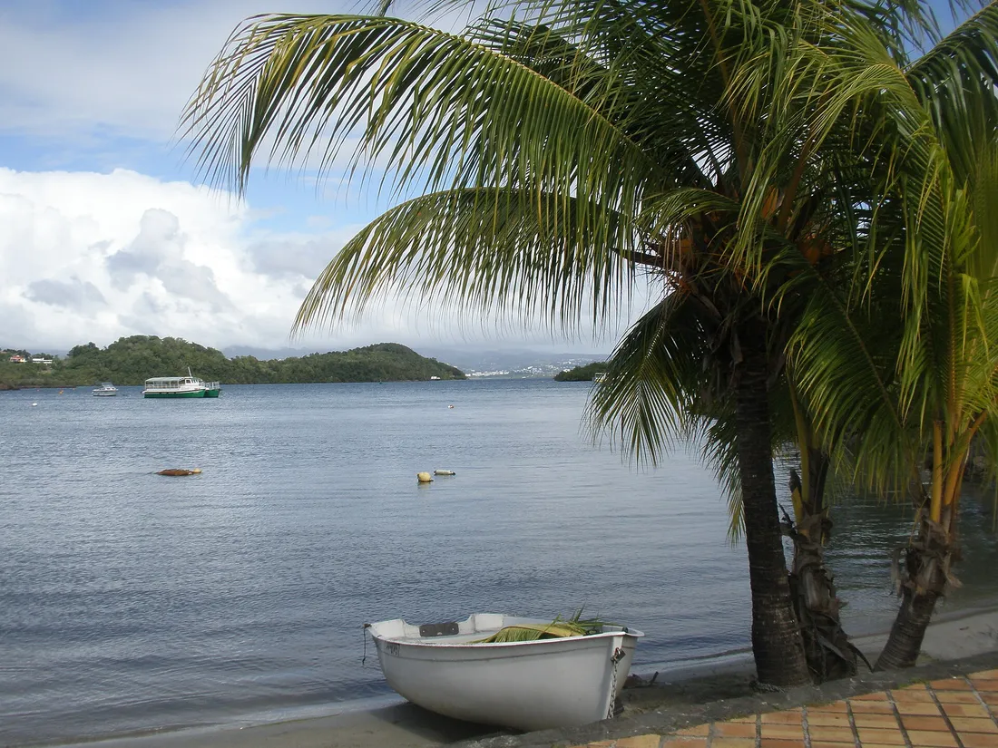 Front de mer du bourg des Trois-Îlets en Martinique, avec une barque échouée, un cocotier et la navette maritime traversant la baie de Fort-de-France en arrière-plan