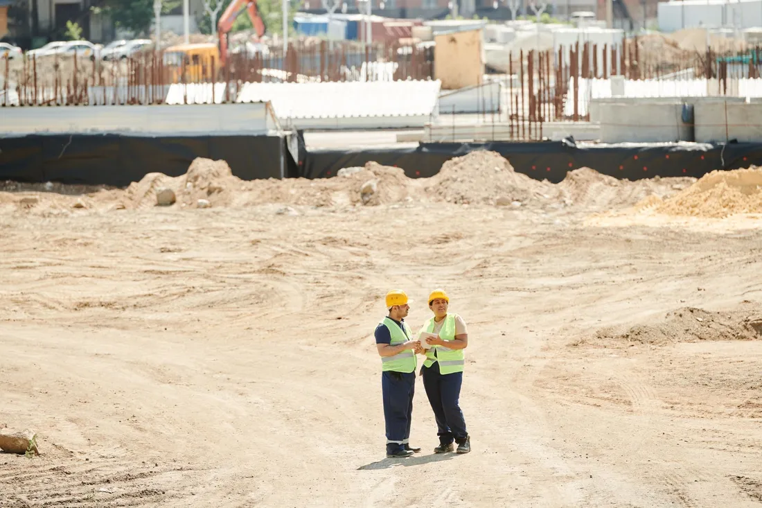 Deux ouvriers du bâtiment en gilet fluorescent et casque de chantier consultent des plans sur un vaste chantier de terrassement