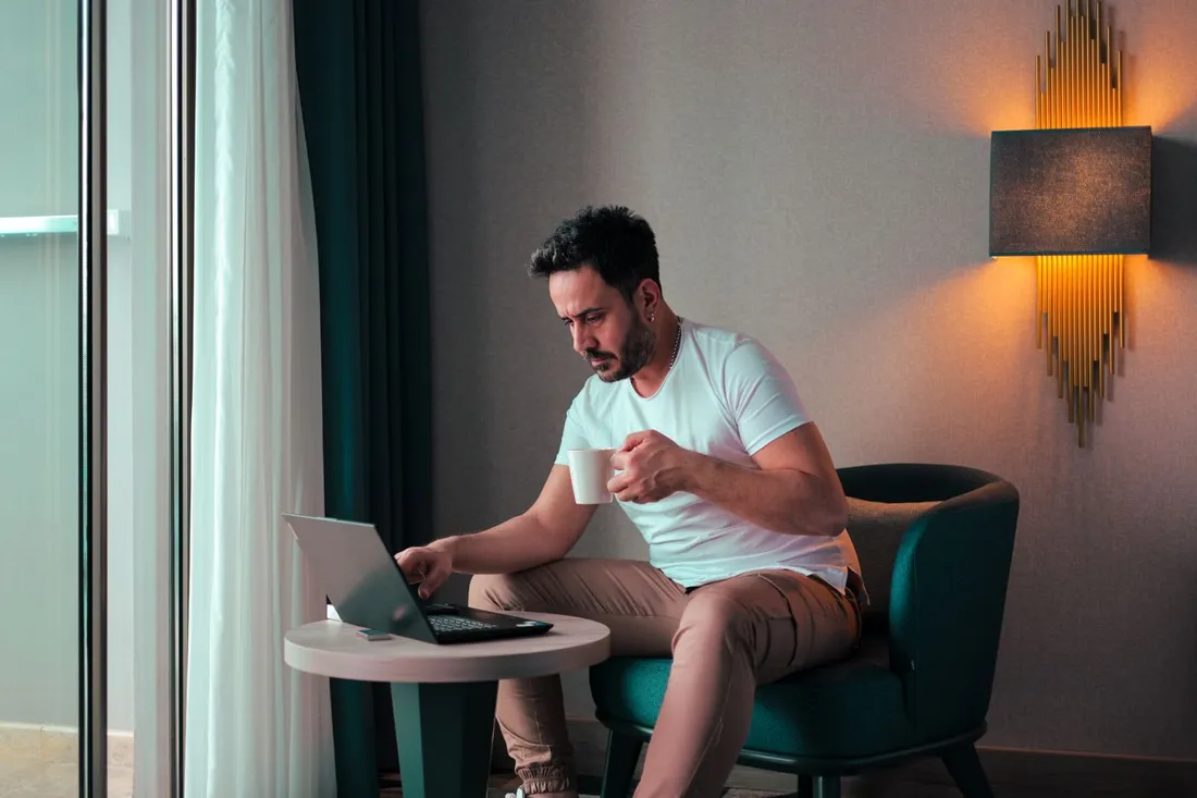Homme en tenue décontractée travaillant sur son ordinateur portable, un café à la main, installé dans un fauteuil près de la fenêtre d'un logement lumineux