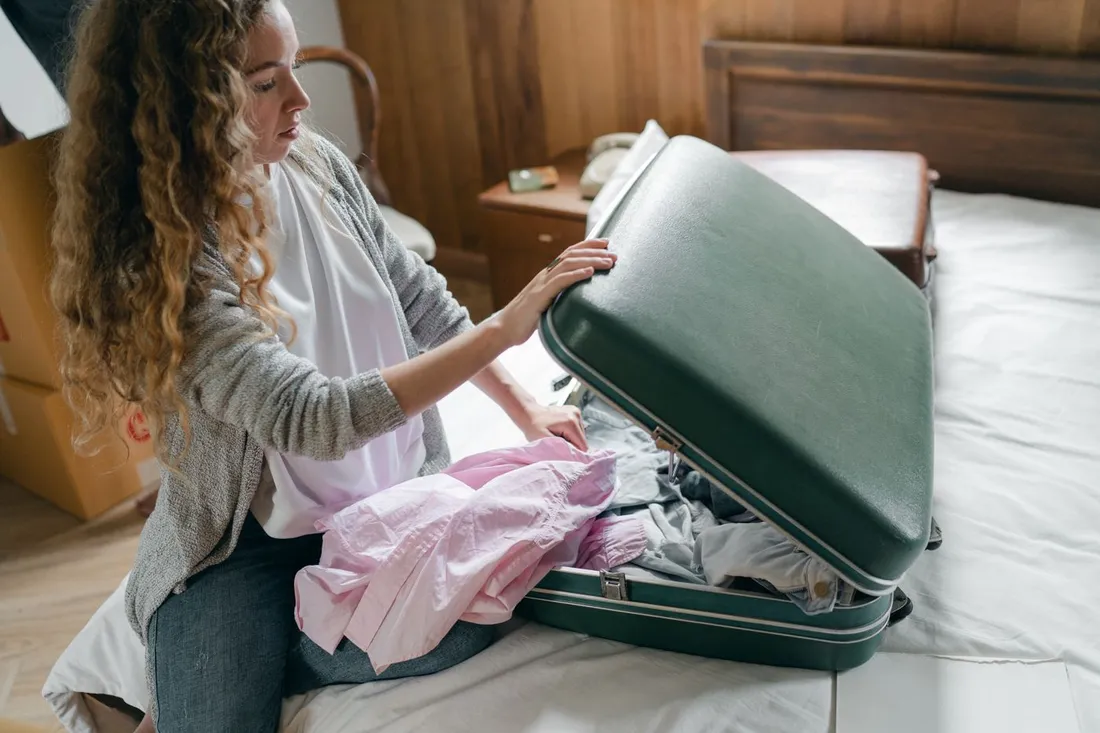 Femme assise sur un lit en train de preparer sa valise ouverte, cartons de demenagement visibles derriere elle