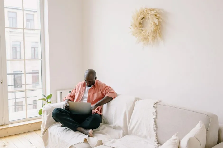 Homme installé sur le canapé du salon lumineux d'un appartement meublé, travaillant sur son ordinateur portable près de la fenêtre