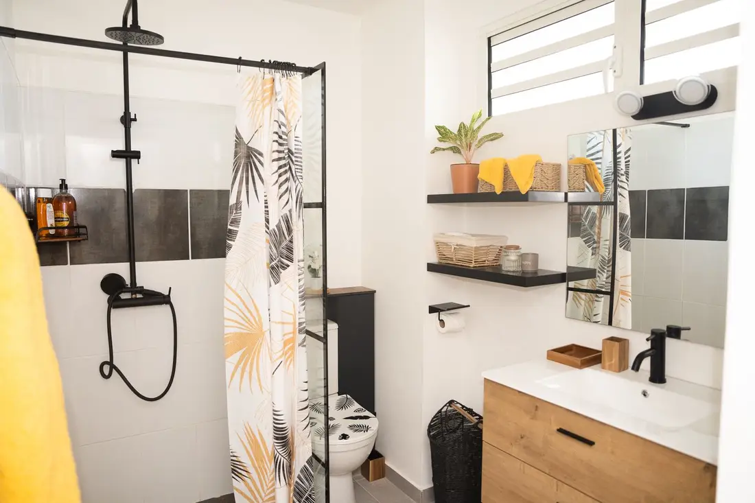 Salle de bain du Cocon Chic : douche à robinetterie noire, rideau à motifs de palmes, meuble vasque en bois clair