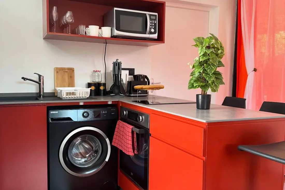 Cuisine rouge d’un appartement de la Résidence Francis : lave-linge et four noirs encastrés, micro-ondes, plan de travail gris avec évier