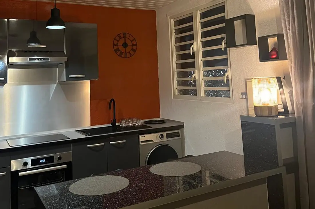 Cuisine d’un appartement Cayenne Chic le soir : mur orange, hotte inox, four encastré, lave-linge et bar en pierre noire