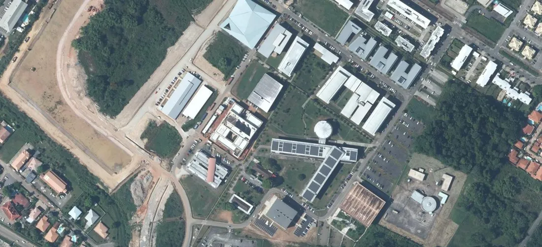 Vue aérienne verticale du campus de Troubiran à Cayenne : bâtiments universitaires à toitures claires, amphithéâtre à toit pyramidal, résidences en peigne, parkings et parcelles boisées de part et d'autre de la route de Baduel