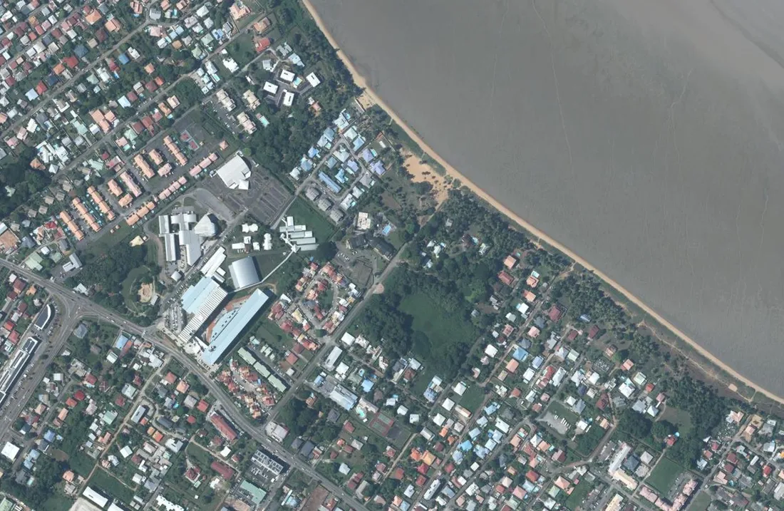 Orthophotographie aérienne verticale de la plage de Montabo à Cayenne : le cordon de sable longeant l'Atlantique et, derrière lui, le quartier Zéphir avec ses maisons à toits clairs et ses équipements publics