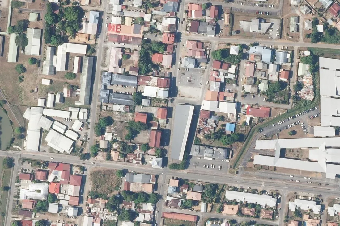 Vue aérienne verticale du centre-ville de Kourou, en Guyane, autour de l'avenue du Général de Gaulle et de la rue Edjide Duchesne : petits immeubles, maisons en tôle et hangars le long des voies