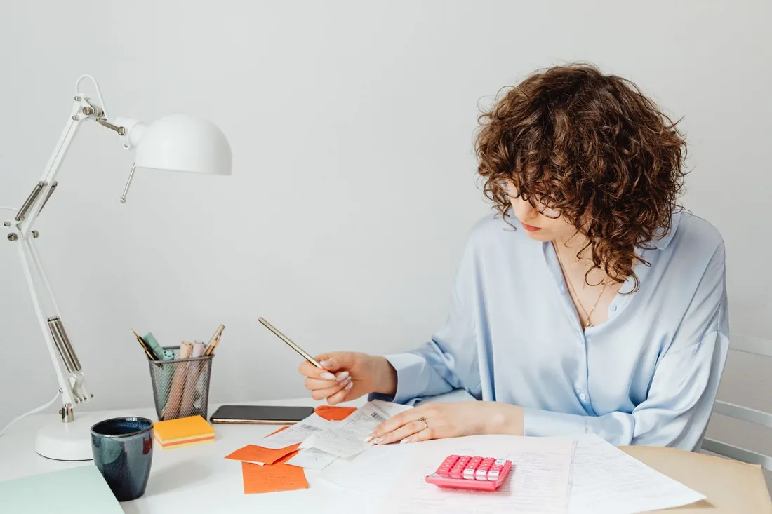 Personne assise à un bureau vérifiant des reçus et des factures avec une calculatrice et des documents, pour préparer une note de frais
