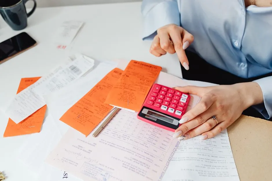 Personne classant des reçus et des factures sur un bureau, calculatrice en main, pour préparer une note de frais de déplacement