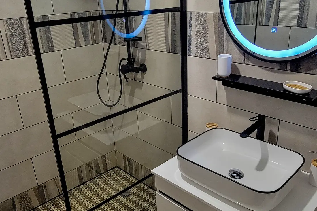 Douche à l’italienne d’un appartement Cayenne Chic : verrière noire, robinetterie noire et miroir rond à éclairage intégré