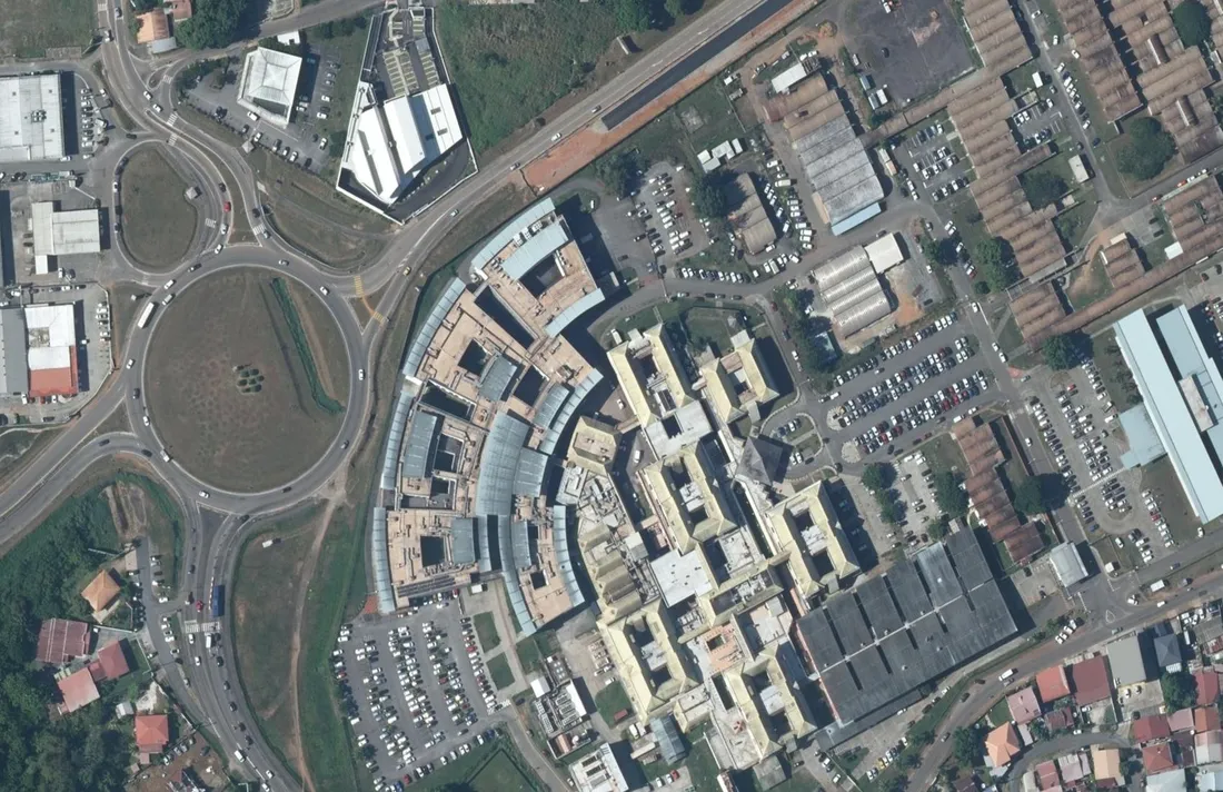 Vue aérienne verticale du CHU de Guyane, site Andrée-Rosemon à Cayenne : le bâtiment en arc de cercle aux verrières, les pavillons à toiture jaune, les grands parkings de personnel et le rond-point Catayée sur la gauche