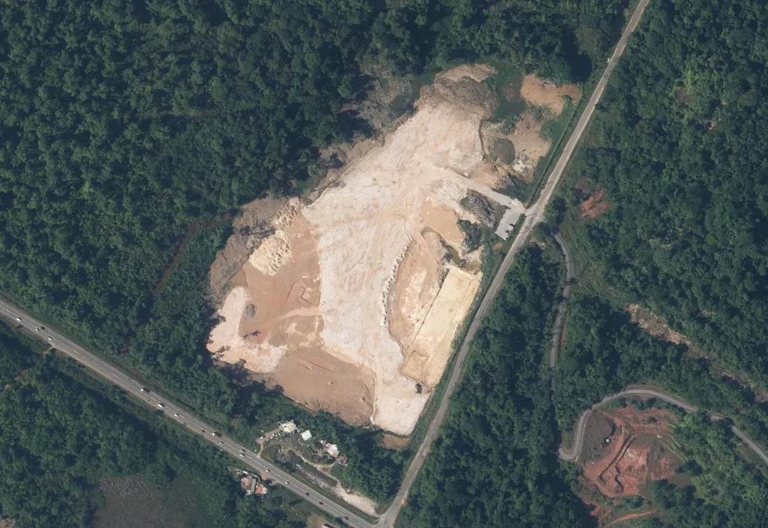 Vue aérienne du site de la centrale du Larivot à Matoury : une vaste plateforme terrassée en terre claire ouverte dans la forêt, la RN1 en bas à gauche et une route départementale le long de la lisière à droite
