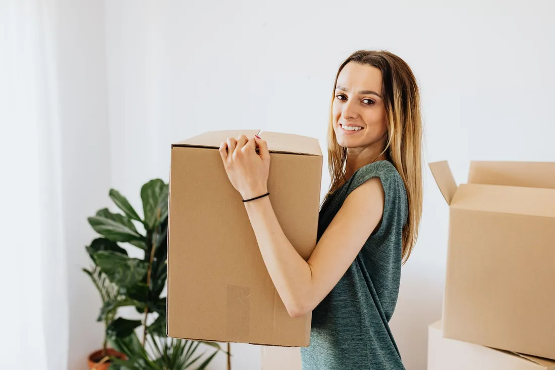 Femme souriante portant un carton de déménagement dans un logement lumineux