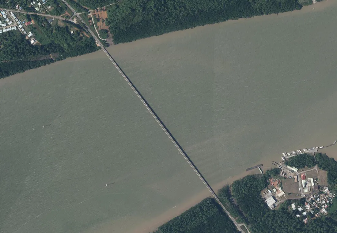 Vue aérienne du pont du Larivot, long ouvrage rectiligne franchissant la rivière de Cayenne en diagonale, avec des véhicules dessus ; berge boisée de Macouria en haut à gauche, port et hameau du Larivot à Matoury en bas à droite