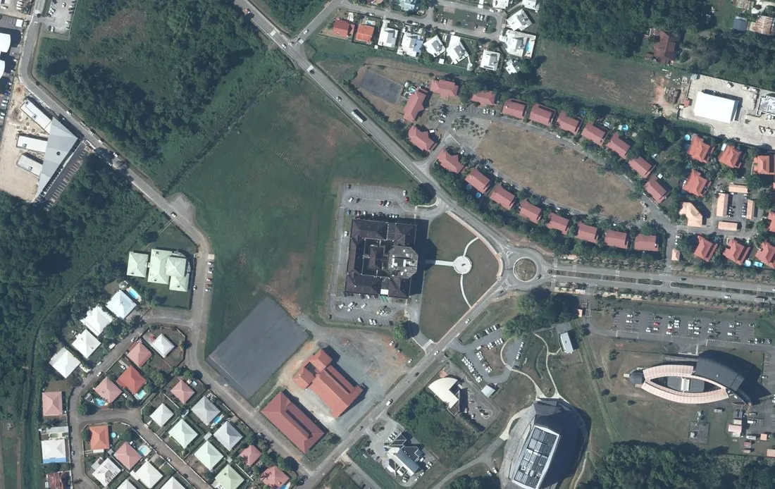 Vue aérienne du centre administratif de Rémire-Montjoly : l'hôtel de ville à toiture sombre au centre, son parvis et son rond-point, entouré de rangées de maisons à toitures rouges et de lotissements pavillonnaires