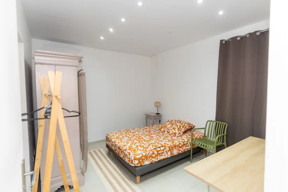 Chambre du Nid de Thalys : lit au couvre-lit orange à motifs, armoire patinée, chaise verte et portant en bois