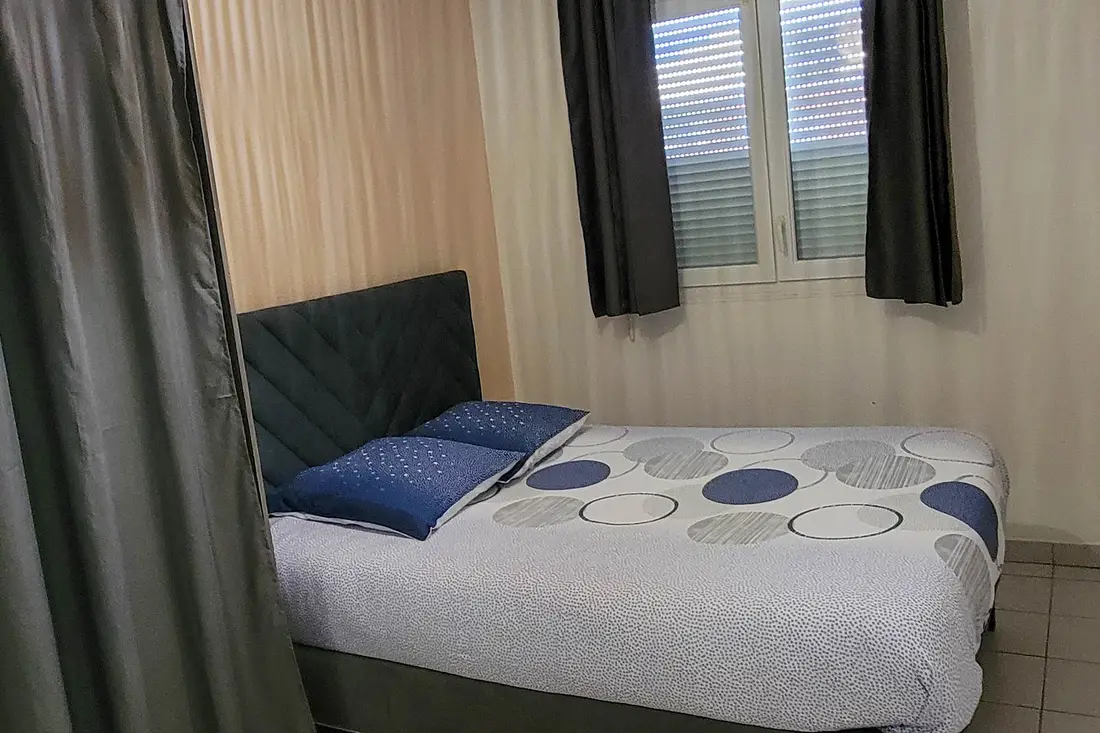 Chambre d’un appartement Cayenne Chic : lit à tête capitonnée sombre, coussins bleus, rideaux gris
