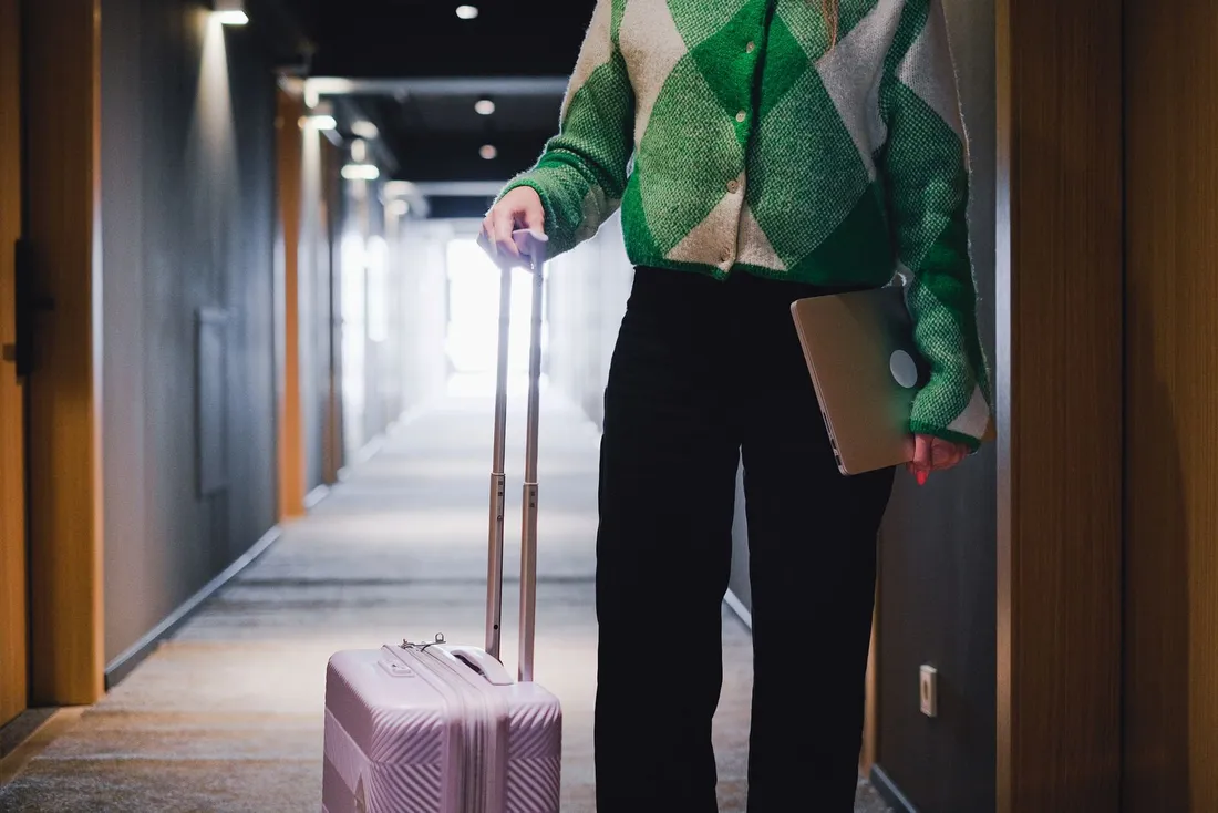 Voyageuse d'affaires tirant une valise cabine et tenant un ordinateur portable dans le couloir d'un hôtel, à l'arrivée dans son hébergement