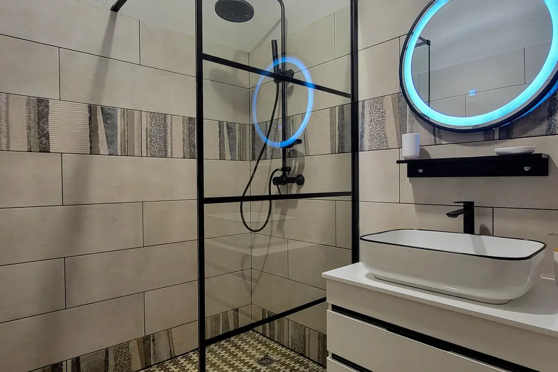 Salle d’eau d’un appartement Cayenne Chic : douche à verrière noire et miroir rond éclairé