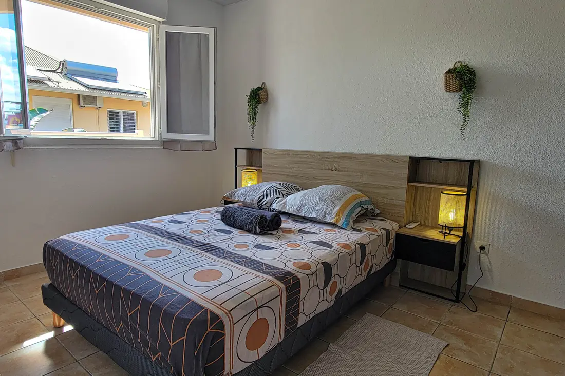 Chambre d’un appartement Cayenne Chic : lit double à tête en bois clair entre deux chevets suspendus éclairés, fenêtre ouverte sur la résidence